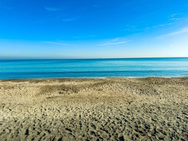 Güneş kumlu bir plajın üzerinde parlıyor, okyanusun uzaklarda uzandığı yerde, açık mavi su ve açık gökyüzü manzarası yaratıyor..