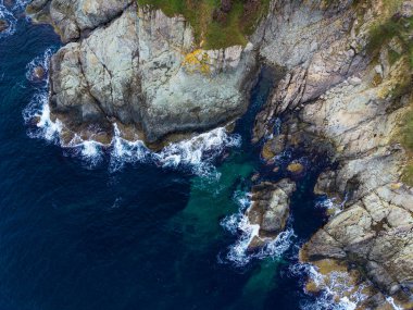 Dalgalar engebeli bir kıyı şeridi boyunca büyük kayalara çarpar. Temiz su yüzeyin altındaki taşları ortaya çıkarır. Gün boyunca doğanın güzelliğini gösterir..