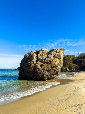 Sakin suyun yanında kumlu bir plajda büyük bir kaya oturur. Gökyüzü açık ve mavi. Ağaçlar uzaktan görülebilir. Gündüz oldu ve güneş parlıyor..