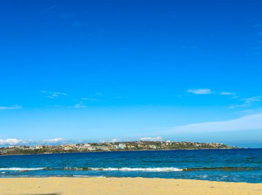Parlak mavi bir gökyüzünün altında kumlu bir plajın geniş bir manzarası okyanusla buluşur. Uzak sahil bazı evler ve yeşillikler gösteriyor. Öğleden sonra dalgalar yavaşça kıyıya vurur..