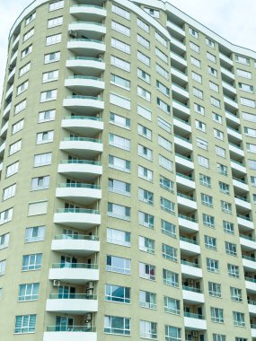 Kentsel bir alanda yüksek bir binada çok sayıda balkon ve çok sayıda pencere bulunur. Tasarım modern mimari tarzını gösteriyor ve gökyüzünde bulut örtüsünü yansıtıyor..