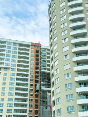 Yüksek binalar yan yana durur. Bir binanın inşaat için yan tarafında iskelesi var. Gökyüzü parçalı bulutlu ve alan işlerle meşgul görünüyor..