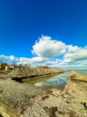 Kumlu bir sahil manzarası ve gökyüzünü yansıtan bir su. Solda bir inşaat alanı var. Görünürde bir bina ve makine var..