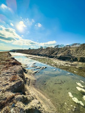 Su, kayalık bir bankanın yanındaki dar bir kanaldan akıyor. Bulutlar yüzeye doğru sürüklenirken güneş ışığı da yüzeye yansıyor. Sahne, açık bir öğleden sonra sahili gösteriyor..