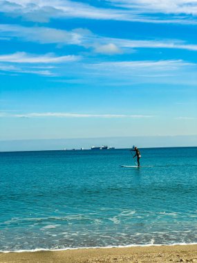 Bir insan sahile yakın temiz suda kürek çekiyor. Kum plajı görünür ve ufukta gemiler var. Gökyüzü açık mavi ve bulutlu.