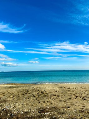 Dalgalar sahildeki kumlu sahile hafifçe dokunur. Açık gökyüzü bulutları ve mavi renkleri gösterir. Uzak karalar suyun ötesinden görülebilir. Birçok insan kıyıya yakın dinlenir..