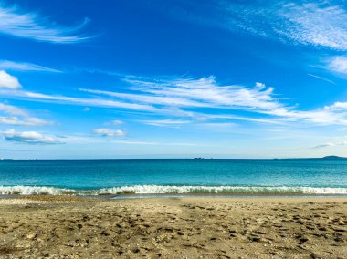 Plaj sahnesinde sahile vuran sakin dalgalara yol açan ayak izleri olan ıslak kumlar görülüyor. Gökyüzü açık ve bulutlar suyun üzerine dağılmış..