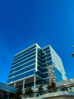 Camdan pencereleri gökyüzüne doğru uzanan uzun bir bina. Yapının birkaç katı var ve iyi tasarlanmış görünüyor. Ağaçlar açık bir alanda, güneşli bir manzara göstererek büyür..