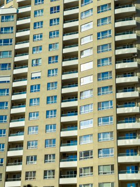 Manzara birçok pencere ve balkonlu yüksek bir apartmanın birden fazla katını gösteriyor. Güneş ışığı dış yüzeyde parlıyor, camda yansımalar yaratıyor..