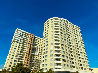 Kentsel bir bölgede mavi gökyüzüne yükselen iki yüksek bina. Güneş parlıyor, mimariyi aydınlatıyor ve çevresindeki yeşillikleri aydınlatıyor..