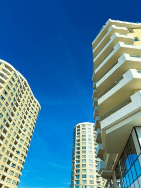 Uzun binalar, görünürde bulutlar olmayan mavi bir gökyüzüne karşı yüksekte dururlar. Yapıların beyaz balkonları ve pek çok penceresi var. Bu da şehir merkezindeki güneş ışığını yansıtıyor..