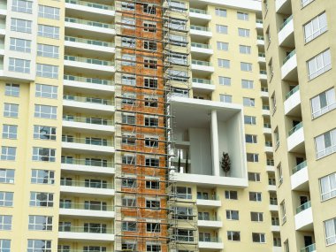 İşçiler yan tarafında iskele olan yüksek bir konut inşa ediyorlar. Açığa çıkmış tuğla ve geniş bir açıklık yapıyı belirler. Sahne gündüz çekiliyor..