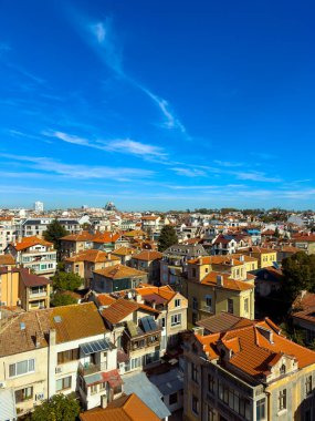 Olay yeri, birçok evi olan bir yerleşim bölgesindeki çeşitli binaları ve çatıları gösteriyor. Gökyüzü açık ve mavidir. Birkaç bulut görülebilir. Bu resim tipik bir şehir ortamını yakalar.