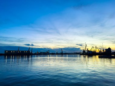 Liman sahnesi, karanlık gökyüzüne karşı vinç ve gemileri gösteriyor. Gün sona ererken, su günbatımının renklerini yansıtıyor. Geceye geçişi işaret ediyor..