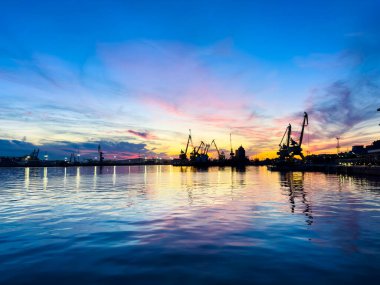 Gün batımı sahnesinde deniz kıyısındaki bir rıhtımda vinçler görülüyor. Işık yüzeye yansıdıkça gökyüzü pembe ve mavi renklere sahiptir. Su turnaların altında dalgalanır.