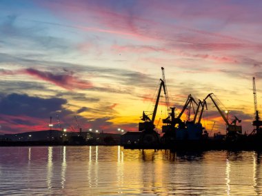 İşçiler rıhtımdaki görevlerini tamamlıyorlar. Vinçler gün batımında renkli gökyüzüne karşı dik duruyorlar. Su, akşam ışığının canlı renklerini yansıtır..