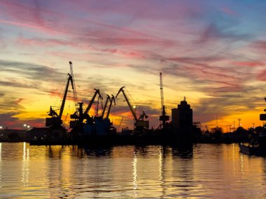 Bir limandaki vinç ve yapılar renkli bir günbatımına karşı siluetlidir. Bulutların ve turnaların yansıması su yüzeyinde görülebilir. Akşam gökyüzü canlı renkler gösterir.