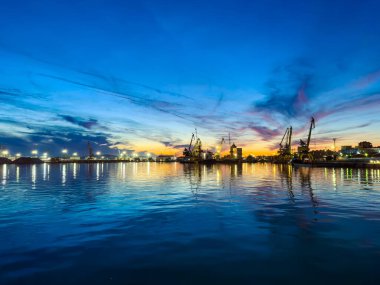 Gün batımında limanda tekneler ve turnalar görülebilir. Gökyüzü mavi ve turuncunun tonlarını gösteriyor. Su, vinçlerden ve etrafındaki binalardan gelen ışığı yansıtır..