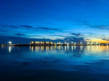 Akşam, suyun binaların ve teknelerin ışıklarını yansıttığı bir limanda. Güneş batarken bulutlar gökyüzünde beliriyor, renkli bir sahne yaratıyor..