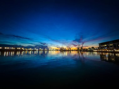Güneş batarken tekneler ve turnalar limanda görülebilir. Gökyüzü mavi ve turuncu tonlarıyla renk değiştirirken ışıklar suyu yansıtıyor..