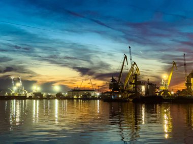 Vinçler ve gemiler güneş batarken bir limanda görülebilir. Gökyüzünde bulutlar var ve su günbatımının renklerini yansıtıyor. Gemilerin ışıkları sahneyi aydınlatıyor..