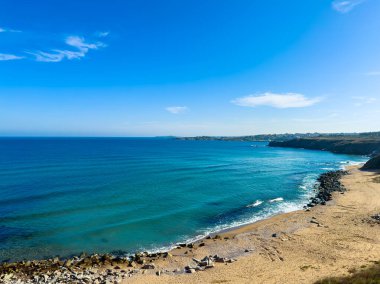 Açık mavi su, parlak bir gökyüzünün altında kumlu plajla buluşur. Güneşli bir günde dalgalar kıyıya vurur kıyılarda, kayalar kıyıları kaplar..