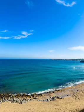 Dalgalar kumlu bir plaja yuvarlanırken kayalar kıyıya vuruyor. Gökyüzü az bulutla parlak ve mavidir. Uzak kara ufukta görünüyor..
