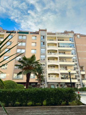 Vatandaşlar, öğleden sonra bulutlu bir gökyüzünün altında yeşil bitkiler ve palmiyelerle çevrili balkonların bulunduğu yüksek bir binanın yanında yürürler..