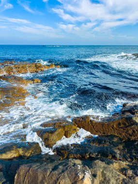 Dalgalar geliyor ve kıyıdaki kayalık yüzeylere çarpıyor. Mavi okyanus berrak bir gökyüzünün altında uzanır..