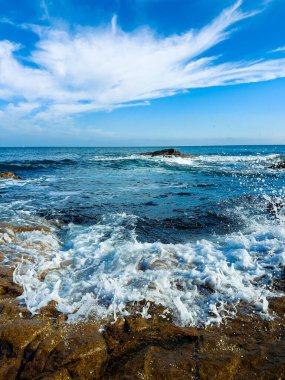 Kıyı şeridinde su taşlara çarpar bulutlar gökyüzünde sürüklenirken. Sahne denizin hareketini ve engin mavi ufku gösteriyor..