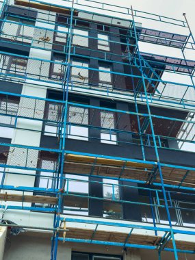 İşçiler binanın üst katlarına ulaşmak için iskeleyi kullanıyorlar. Yapı, mavi çerçeveli ve ağ örgülü devam eden yapı işaretlerini gösteriyor..