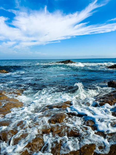 Güneş ışıl ışıl parlarken dalgalar kıyı şeridinde kayaların üzerinde yuvarlanıyor. Deniz, gün ortasında gökyüzünde birkaç bulutla çok uzaklara doğru uzanıyor..