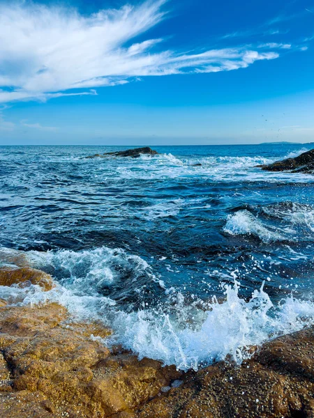 Dalgalar kıyıdaki kayalara vurur ve güneş doğarken su sıçratır. Gökyüzü bulutlarla parlıyor. Su ufka kadar uzanıyor..