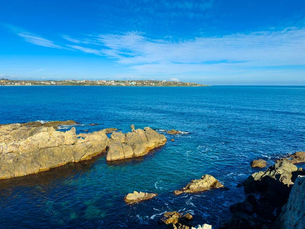 Parlak mavi su, kayalık oluşumlar ön planda dururken berrak gökyüzünün altında güneş ışığını yansıtır. Sahil şeridi ufuk boyunca uzanır ve arka planda binalar vardır..