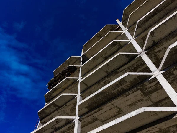 Beton yapı, tamamlanmamış bir tasarım ve açık mavi gökyüzü ile ayakta duruyor. Çelik kirişler görünür durumda. İnşaat alanında çalışmalar devam ediyor..