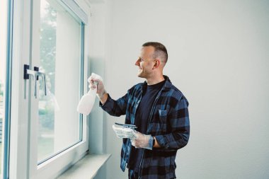 Gündüz vakti parlak bir odada pencereleri temizleyen bir kişi var. Eldiven ve ekose gömlek giyiyorlar. Bir elinde sprey şişesi, diğer elinde temizlik bezi tutuyorlar..