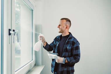 Biri iyi aydınlatılmış bir odada pencereyi temizlemek için sprey ve bez kullanıyor. Bireyler bu ev işi üzerinde çalışırken gülümsüyor..