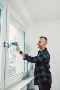 Beyaz duvarlı bir odada bir adam pencereyi temizliyor. O, işi yaparken bir cam sileceği tutuyor ve gülümsüyor. Yakınlarda bir şişe temizlik solüsyonu duruyor..