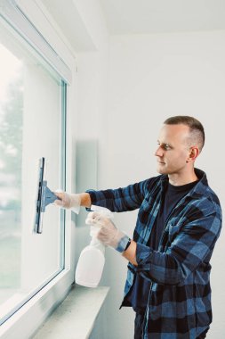 Bir adam pencereyi temizlemek için cam sileceği kullanır. Eldiven takıyor ve elinde bir sprey şişesi tutuyor. O, bardağı netleştirmeye odaklandı. Ayarlar parlak ve basit..
