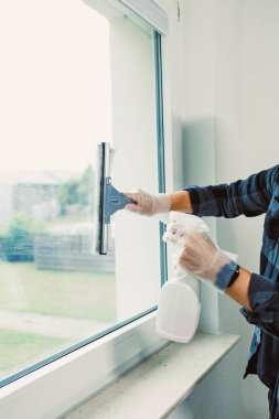 Eldiven giyen bir kişi, kapalı bir alanda sprey şişeyi tutarken pencereyi temizlemek için cam sileceği kullanır. Temizlik yapılırken güneş ışığı camdan içeri giriyor..