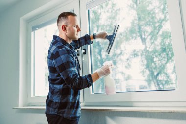 Adamın biri pencereye temizleyici sıkar ve silip temizlemek için cam sileceği kullanır. Güneş ışığı camdan içeri giriyor ve kapalı alanı aydınlatıyor. Korunmak için eldiven giyiyor..