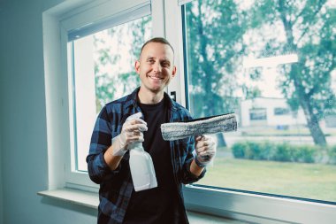 Elinde bigudi ve sprey şişesi olan bir adam pencerenin yanında duruyor. O bir evin camını temizlerken güneş ışığı pencereden içeri giriyor..