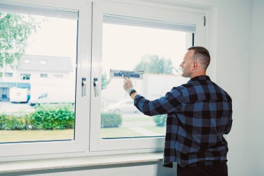 Bir kişi bir evin içinde duruyor ve bir cam sileceği ile pencereyi temizliyor. Manzara bulutlu bir günde bahçeyi ve evleri gösteriyor. Görev sırasında eldiven takan kişi.