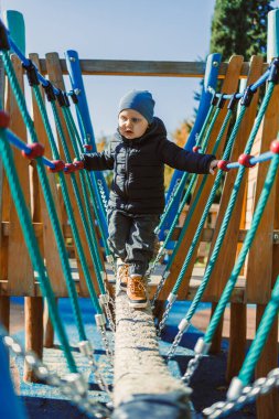 Bir çocuk sonbahar boyunca bir oyun parkında ip köprüde dikkatle yürür. Sahne, çocuk oynarken dengeye odaklanırken arka planda renkli ağaçları gösteriyor.