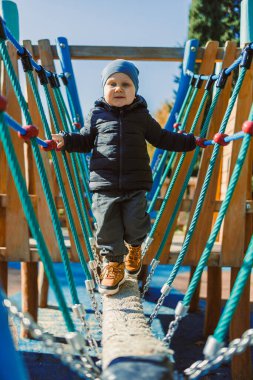 Küçük bir çocuk oyun parkındaki ip köprüde dikkatle yürür. Mavi gökyüzü olan güneşli bir sonbahar günü. Çocuk sıcak bir ceket ve şapka giyiyor, mutlu ve odaklanmış görünüyor..
