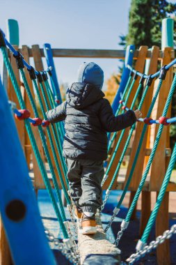Küçük bir çocuk oyun parkında denge kirişinde ipleri destek olarak kullanıyor. Sonbahar geldi ve güneş parlıyor. Ağaçlar arka planda görünür..