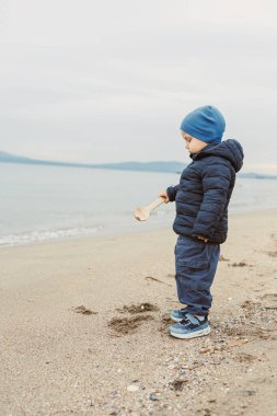 Bir çocuk suya yakın kumda duruyor. Tahta bir kaşık tutuyor ve denize bakıyor. Gökyüzü gri ve atmosfer sessiz..