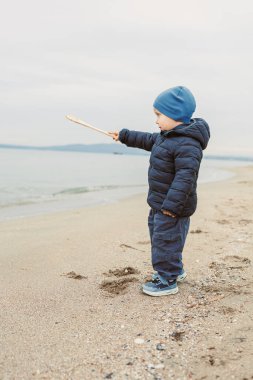 Bir çocuk mavi bir şapka ve ceket giyiyor, sahilde duruyor ve sopayı suya doğrultuyor. Gökyüzü bulutlu ve çocuk ilerideki suya odaklanmış durumda..