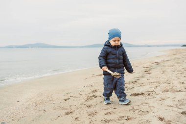 Bir çocuk elinde sopayla kumsalda yürüyor. Su sakin ve gökyüzü bulutlu. Çocuk serin hava için sıcak giyinmiş..