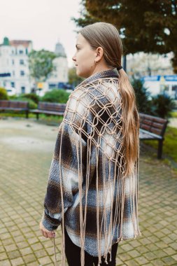 Parkta üzerinde makrome detaylar olan desenli bir bluz olan bir kadın duruyor. Gün boyunca arka planda görünen bir şehir manzarası ile kameradan uzağa bakıyor..